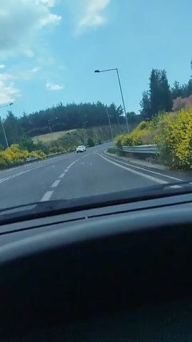 The video shows a view from inside a car looking out at a highway. The highway is curved and surrounded by green trees and yellow bushes. There are streetlights visible along the side of the road. A white car is driving on the highway in the distance. The sky is blue with some clouds. The interior of the car, including the dashboard, is visible in the foreground. The video is shot from a first-person perspective, as if the viewer is the driver.