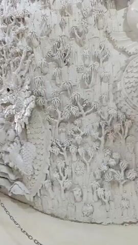 A close-up shot of a detailed white sculpture of a dragon. The dragon appears to be coiled and is intricately carved with scales and facial features. The background of the sculpture depicts a landscape with trees and rocky textures. Small reflective elements, possibly crystals, are embedded throughout the sculpture, catching the light. In the foreground, a silver chain barrier with a gold-capped metal post is visible, indicating a display or restricted area. The lighting is bright, illuminating the white surface of the sculpture.