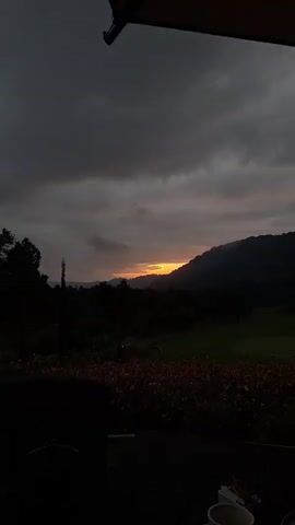 The video opens with a dark, overcast sky, transitioning to a scene of a golf course nestled among rolling hills. The sun is setting, casting a warm orange glow on the horizon, partially obscured by dark clouds. In the foreground, a table is set with a white coffee cup, a saucer, and a plate of food, suggesting a dining or relaxation setting. The overall ambiance is serene, with the natural beauty of the landscape dominating the frame.
