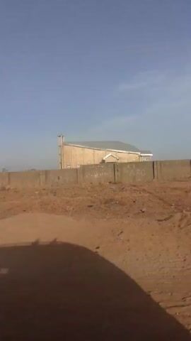 The video shows a tan-colored building, possibly a church, with a silver or grey roof and a bell tower. It is surrounded by a concrete wall. The foreground is a dusty, barren landscape with some dry vegetation. The sky is blue and clear. The camera appears to be moving, possibly from a vehicle.