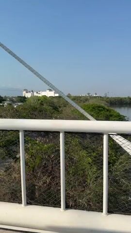 The video shows a view from a bridge with white railings, overlooking a green landscape with a river or body of water. In the distance is a large white building with castle-like towers and a colorful hot air balloon. A road and parking lot are visible near the building. The camera then shifts to show a person wearing sunglasses riding in a golf cart, with the bridge structure visible above. The sky is clear and blue.