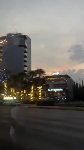 The video depicts a street scene at dusk, likely captured from a moving vehicle. Several buildings are visible, including a tall glass skyscraper with balconies lit by yellow lights and a building labeled "La Encomienda." Palm trees line the street, also adorned with yellow lights. Cars are moving along the road, and a building with a large "A" logo is visible, possibly a bank. There is also a billboard and digital sign displaying an image of food. The sky is a mix of grey and orange, indicating either sunset or sunrise. The overall atmosphere suggests an urban commercial area.