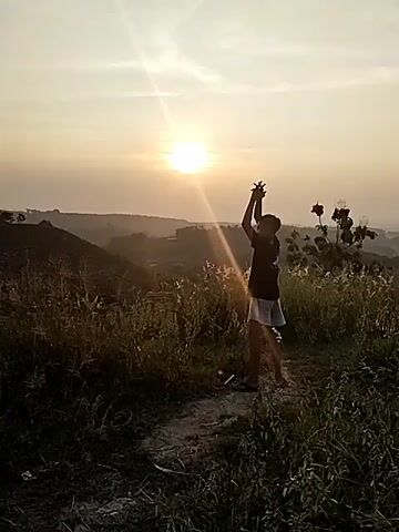 The video features a person, likely male and young, standing on a narrow dirt path amidst a grassy, hilly landscape. The setting is outdoors, possibly on an elevated viewpoint, with a panoramic view of distant hills and a hazy horizon. The time of day is either sunrise or sunset, indicated by the bright, low-hanging sun, which creates a prominent lens flare and casts warm, golden light across the scene. The weather appears clear with no visible clouds.

At the beginning of the video (00:00-00:02), the person is facing partially towards the camera, with both arms raised high above their head, palms open, seemingly embracing or gesturing towards the sun. They are wearing a dark t-shirt and light-colored shorts. The camera then briefly loses focus (00:02-00:03), blurring the image. When it refocuses (00:03-00:05), the person has turned to face away from the camera, looking out at the distant landscape and the sun with their arms lowered. They appear relaxed, observing the view.

The foreground and midground are covered in tall green and dry grass, with a few sparse plants visible. The overall scene conveys a sense of tranquility and appreciation for nature. No visible text is present in the video. The location context provided is Jambeyan, Indonesia.