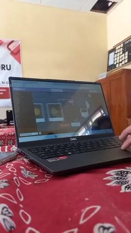 A person's hand is visible on the right side of the frame, resting on the keyboard of a black DAC laptop. The laptop screen displays an online educational platform with interactive questions and images of a fish, a cat, and a robot. The laptop is placed on a red cloth with a white floral pattern. In the background, a yellow wall is visible, with a white banner that has red lettering and the word "GURU" prominently displayed. To the right of the banner, there is a wooden cabinet and a bulletin board with papers pinned to it. The lighting suggests an indoor setting, possibly a classroom or office.