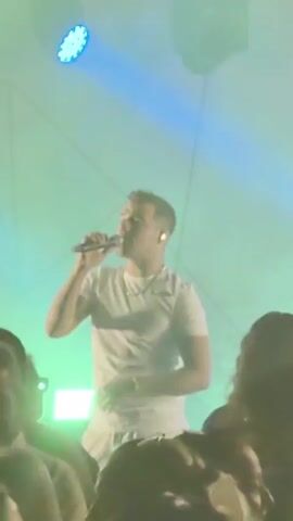 A man in a white t-shirt sings into a microphone on a stage. He has his left hand on his stomach. He is wearing a necklace and a bracelet. There is another person on the stage behind him holding a guitar. The stage is lit with green and blue lights. There is smoke on the stage. A crowd of people are visible in the foreground, silhouetted against the stage lights.