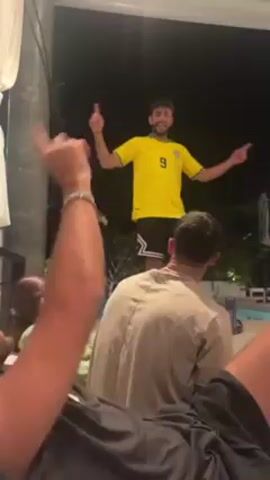 The video shows a man in a yellow Brazilian soccer jersey dancing with his arms outstretched near a pool at night. Two people lounge on a patio set, watching the man dance. A woman is also seen sitting on the patio, smiling. The scene appears to be a casual, relaxed gathering outdoors. The background features greenery and a dimly lit area.
