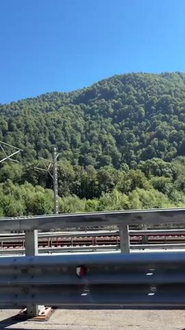 The video shows a view from a moving vehicle, likely a car or train, on a bright, sunny day. The foreground features a metallic guardrail. In the middle ground, a train track with a red train is visible. Beyond the tracks, a lush, green forested mountain rises against a clear blue sky. Several utility poles with wires are also present.