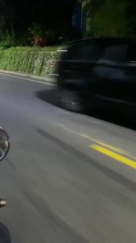 This video shows a point-of-view perspective from a vehicle, likely a motorcycle, driving at night on a paved road. The road has a solid yellow line on the right side and a white line on the left. Lush green foliage lines the side of the road, with some bushes and trees visible. In the distance, there's a low stone wall adorned with colorful mosaic tiles, and a vintage-style lamppost illuminates the greenery.

At one point, a person on a bicycle is seen near the edge of the road, and another motorcycle passes by. The overall scene suggests a tropical or subtropical location, given the dense vegetation. The time of day is clearly night, with artificial lighting from the lamppost and vehicle headlights, although the specific weather conditions are not apparent. The setting appears to be a road in or near a residential area or a scenic route, possibly in Madiun, East Java. There is no visible text in the video. The motion blur indicates the vehicle is in motion.