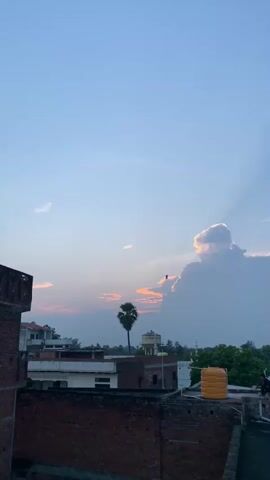 The video is a vertical shot of a sunset scene. The sky is a pale blue with some scattered white clouds and darker clouds forming a large mass on the right, lit from behind by the setting sun, creating crepuscular rays. In the foreground, there are rooftops of buildings, some made of red brick and others with tiled roofs. A person wearing dark clothing is sitting on the edge of a rooftop. There is a large yellow water tank on one of the rooftops. A single palm tree stands in the distance, and other trees and buildings are visible on the horizon. The overall atmosphere is calm and serene.