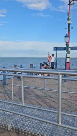 The video shows a pier by the sea under a blue sky with scattered clouds. Two people are on the pier. One person, wearing a dark shirt and shorts, is crouched down, appearing to be tending to something on the ground. Another person, wearing a white shirt and dark pants, is standing and holding a fishing rod. There is a life preserver attached to a pole nearby. The pier has a metal railing in the foreground.