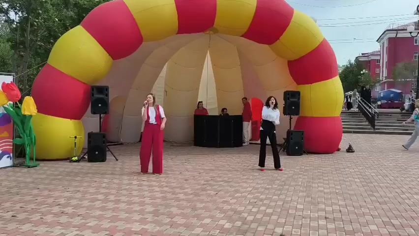 Two young women, one in red overalls and white shirt, the other in black pants and white shirt, are singing into microphones in front of a large inflatable archway. The archway is red and yellow striped. Behind them, several people are visible, including some near a DJ booth. Speakers are placed on either side of the stage area. There are large inflatable tulips to the left of the archway. The scene appears to be an outdoor public space with buildings in the background and people walking in the distance.