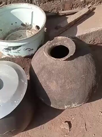 The camera pans around a collection of used household items placed outdoors. There's a large, rounded clay pot with a broken rim, a metal pot with a lid, a blue enamel pot, a black plastic bag, some pieces of wood, a Coca-Cola crate with bottles, and other debris. The ground appears to be dirt or sand. The items are sitting against a wall or foundation.
