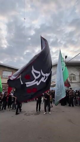 A group of people are gathered in a street in Babol, Iran, holding and waving large flags. The flags are varied in color, with prominent black, white, and green flags being held by individuals. The sky above is cloudy, suggesting it is either dawn or dusk. Buildings line the sides of the street, some appearing to be residential homes with windows and balconies, while others seem to be part of a more commercial or public area, possibly a mosque with an arched entryway. The people in the foreground are holding the flags on poles, with some individuals appearing to be actively participating in a procession or ceremony. Many of the participants are wearing dark clothing, which could indicate mourning or a religious observance. The overall atmosphere seems solemn or celebratory, depending on the nature of the event. The scene is likely taking place outdoors, in a populated urban or semi-urban environment. There is a string or wire visible in the upper portion of the frame, suggesting the presence of utility lines or temporary decorations. The flags themselves are decorative and symbolic, and the act of waving them implies a collective expression or demonstration. The presence of multiple flags and a gathering of people suggests a significant event is occurring, potentially a religious procession or a cultural festival.