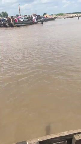 The video shows a wide, muddy river under a cloudy sky. On the left bank, several boats are docked or approaching. One boat appears to be a ferry carrying passengers and a white car. People are visible on this boat and on the bank. Other boats, some appearing to be smaller and loaded with goods, are further out in the river. The right bank is distant and appears to be a sandy or rocky area with some vegetation. The water is rippled, indicating a gentle current. The overall scene depicts a busy river crossing or port.