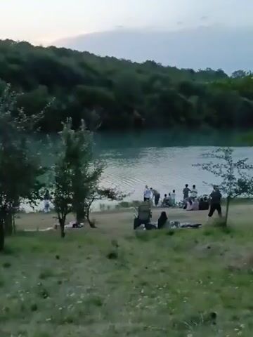 The video shows a wide shot of a body of water, likely a lake or quarry, with a wooded hillside in the background. In the foreground, there is a grassy area with several trees and bushes. A group of people are visible on the grassy bank near the water. Some are standing, and some are sitting on the ground or in chairs. There are also some picnic blankets spread out on the grass. The overall impression is of a leisurely outdoor gathering.
