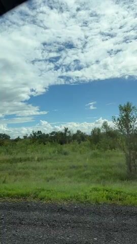 The video shows a view from inside a vehicle driving on a paved road. The scenery outside consists of green fields and trees under a partly cloudy sky. The camera captures the road ahead and the surrounding landscape.