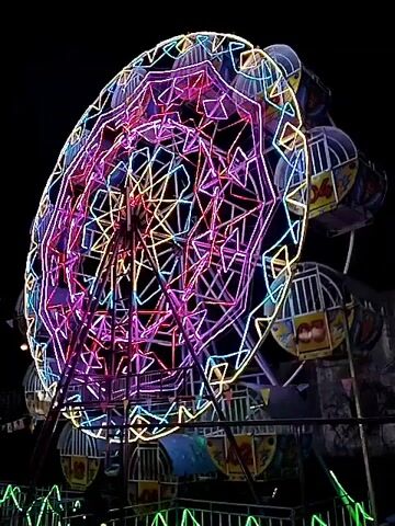 The video shows a Ferris wheel at night. The wheel is lit up with colorful neon lights. The cabins are painted with different numbers. The wheel is spinning slowly. The background is dark.