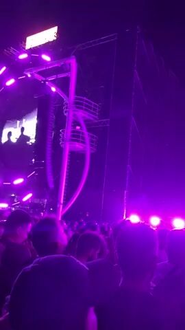 A concert crowd is visible, illuminated by purple stage lights. The stage itself is largely dark, with a large screen that is also dark. There are two figures on the stage, one possibly holding a microphone. The overall atmosphere is that of a live music event.