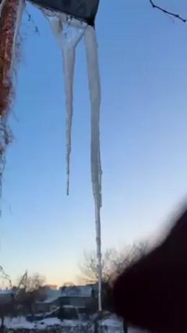 A hand detaches a large icicle hanging from a metal eave against a clear blue sky. Two icicles are visible, one much larger than the other. The hand snaps off the larger icicle. The hand then holds the icicle, moving it against the sky, and then drops it. Snow-covered rooftops are visible in the background. The scene appears to be an outdoor winter setting.