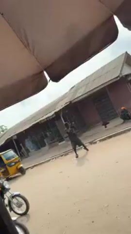 The video shows a street scene, apparently in a West African country, with a dusty road and a row of buildings with corrugated metal roofs. Under a large umbrella, the camera records a man on a motorcycle and a yellow auto rickshaw. In the street, a Black man in a black outfit appears to be in distress, flailing his arms and stumbling, seemingly disoriented. Another man in a purple and yellow patterned shirt is walking towards him. Other figures are present in the background, one kneeling in front of a building. The video is shot from an elevated position, partially obscured by the umbrella.
