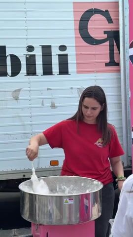 A young woman stands in front of a cotton candy machine, making cotton candy. She is wearing a red t-shirt and dark pants. The cotton candy machine is pink and has a silver bowl. Behind her is a truck. A banner with a design is on the far right.