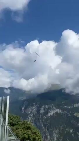 A paraglider is soaring in the sky above a mountainous landscape.  The sky is a vivid blue, dotted with fluffy white clouds. Below, the mountains are covered in dense, green forest. The camera angle shifts to include a building with a glass railing and people sitting at tables in the foreground, suggesting a restaurant or cafe.