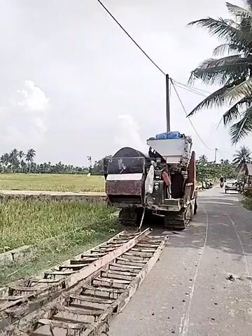 The video features a large, tracked combine harvester parked on the side of a paved road in Dayahbaro, Indonesia. The machine, which appears aged and industrial, has a white hopper-like structure on top and a long, wooden ramp extending from its rear onto the road. The setting is rural, with expansive green rice fields bordering the road. Palm trees are visible in the distance under a bright, partly cloudy sky. A utility pole with power lines runs alongside the road. The combine harvester is stationary throughout the video, and no people are clearly visible interacting with it. There is no clearly legible text on the machine.
