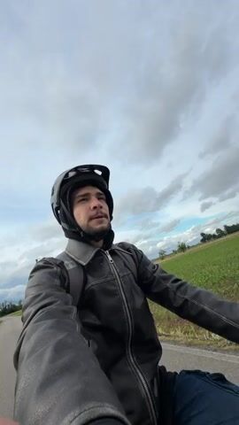 A man wearing a black leather jacket, a black backpack, and a black helmet is riding a bicycle on a paved road. He is looking directly at the camera, then shifts his gaze slightly to his right. The background consists of a field of green crops and a cloudy sky. Power lines are visible on one side of the road. The lighting suggests an overcast day. The camera appears to be mounted to the bicycle's handlebars or held by the cyclist, providing a first-person perspective of the ride.
