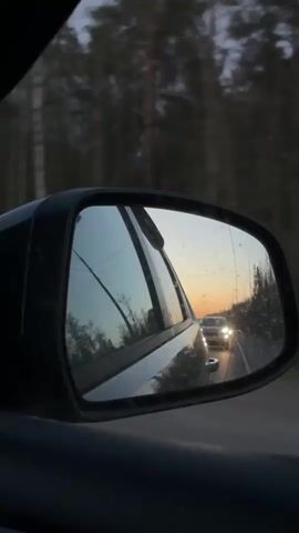 The video shows the side view mirror of a car.  The sky in the mirror is lit with a dim orange light from a sunset or sunrise, with light blue visible above.  A car with headlights on is visible further down the road, as well as trees on the side of the road.  It appears the camera is in a moving car.