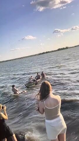 Two young women stand in shallow water on the left and right sides of the frame. The woman on the left wears a black dress and holds a camera, taking a photo. The woman on the right wears a light-colored tank top and shorts, facing away from the camera. In the middle of the frame, a group of men are in the water, some in a small boat and others in the water. One man is in the water, holding onto the back of the boat, while three men are in the boat, one of whom appears to be rowing or paddling. The water is choppy, and the sky is blue with some clouds. The scene takes place on a sunny day.
