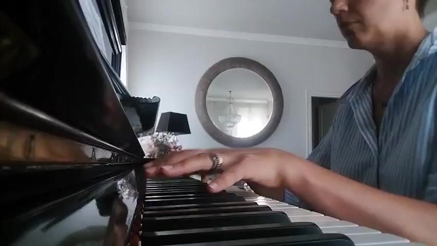 A person is playing a piano. The camera is close up on the piano keys and the person's hands. The person is wearing a striped shirt and rings on their fingers. In the background, a round mirror with a chandelier visible inside it hangs on a gray wall. A black lamp and a bouquet of flowers are on a surface behind the piano. The lighting suggests it is daytime indoors.