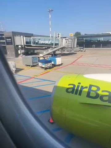 The video shows an airplane engine with "airBaltic" written on it in green letters. The engine is parked at an airport gate. In the background, there is an airport terminal building, a jet bridge, and a control tower. The sky is blue and clear. The video appears to have been taken from inside the airplane.