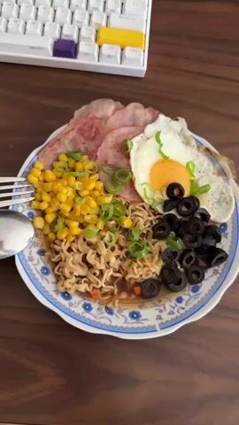 A plate of food sits on a wooden table. The plate contains instant noodles, corn, ham, a fried egg, sliced black olives, and chopped green onions. A fork and spoon are on the left side of the plate. In the background, a portion of a white keyboard with yellow, blue, and purple keys is visible. The video shows a close-up view of the food, with some frames slightly out of focus.