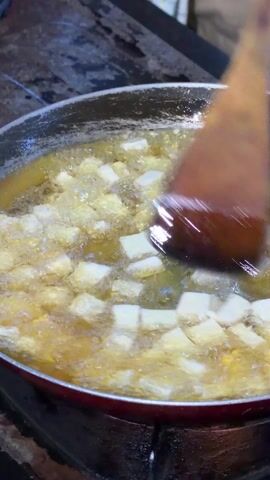 A close-up shot shows small, white, square-shaped tofu pieces frying in a pan filled with bubbling, yellow oil. A brown, wooden spatula is used to stir and turn the tofu. The pan has a red rim and sits on a black stovetop. The background shows a partially visible checkered cloth and a blurred, indistinct area. The camera remains static throughout the video, focusing solely on the frying tofu and the spatula's movement.
