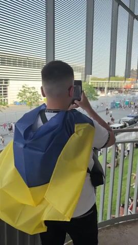 The media file shows a person standing on a balcony, draped in a Ukrainian flag. They are holding up a phone, seemingly taking a picture or recording a video of the scene below. The setting appears to be an outdoor event, possibly a concert or festival, held in front of a large, modern building with a striped facade. Below the balcony, a crowd of people are milling about on paved ground and a grassy slope. Some individuals are sitting on the grass, while others are walking and gathering in groups.

The scene is illuminated by natural light, suggesting it is daytime, likely late afternoon or early evening given the angle of the light. The weather appears pleasant, with people dressed in casual summer attire. The background shows more of the building, trees, and a street with some vehicles. There are barriers and fencing, indicating crowd control measures.

A prominent banner is visible in the distance with text that appears to read "NIVEAHAUS 021" and "PARKPLATZ F+P". The overall atmosphere seems to be one of anticipation and gathering for an event. The presence of the Ukrainian flag suggests a possible demonstration of solidarity or support.