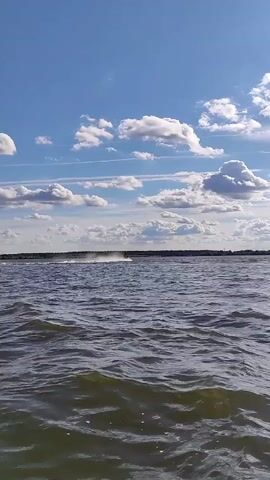A jet ski is speeding across a body of water, leaving a trail of spray. The sky is blue with scattered white clouds. The water appears choppy. The environment is a wide open body of water with a treeline visible in the distance.