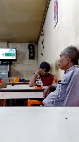 The video shows three men sitting at tables in a small café-like setting. One man is older, wearing a light gray shirt with patterned sleeves, and appears to be talking. A second man, wearing a black hat and a red shirt, is sitting at the same table with a small glass or cup in front of him, and he appears to be listening. At the table to the left, a third man in a black shirt with orange trim is sitting and looking towards the other two. The tables are light-colored and the chairs are orange. There is a television on the wall showing a nature scene, and speakers are mounted on the wall. The walls are painted a light, off-white color. There is a poster of some kind hanging on the wall to the right. The overall lighting is somewhat dim.