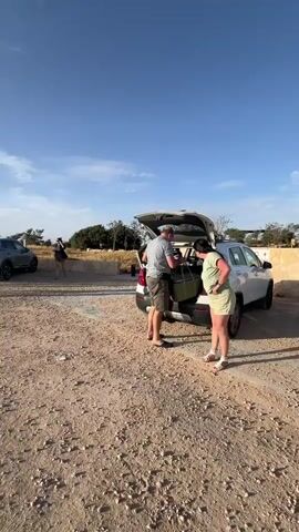 A group of people stand near a wind turbine on a clear day. The video starts with a view of two cars parked on a dirt lot.  Some people are near the back of one of the cars, unloading items.  The camera then pans up to reveal a tall white wind turbine labeled "13". Other wind turbines are visible in the background.  The group of people are standing near the base of the turbine. The sky is blue with some clouds.