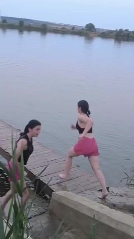 Two young women, one wearing a black sports bra and pink shorts, the other a black and white patterned top and pink shorts, stand on a wooden pier. The first woman runs to the end of the pier and jumps into the water, creating a large splash. The second woman watches her. The pier is made of weathered wood and extends into a calm body of water. Reeds and tall grass are visible in the foreground. The background shows a distant treeline and a hazy sky.