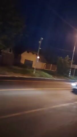 The video shows a nighttime view from a vehicle moving on a road. Several vehicles are visible, including a white car with yellow accents, an orange truck, and a dark-colored car. Traffic lights are present and appear to be green. Streetlights illuminate the scene, casting a warm glow. The environment includes buildings, trees, and utility poles. The footage is somewhat blurry due to the motion and low light conditions.