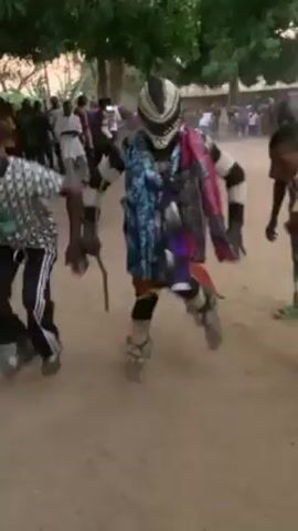 The video shows a person in a traditional costume dancing in a dusty, outdoor environment. The dancer wears a distinctive mask with a black and white pattern, along with colorful patterned cloths draped over their shoulders. They are also carrying a wooden staff. Two other people are dancing alongside the costumed figure, and a crowd of onlookers can be seen in the background. The setting appears to be a village, with trees and simple buildings visible.