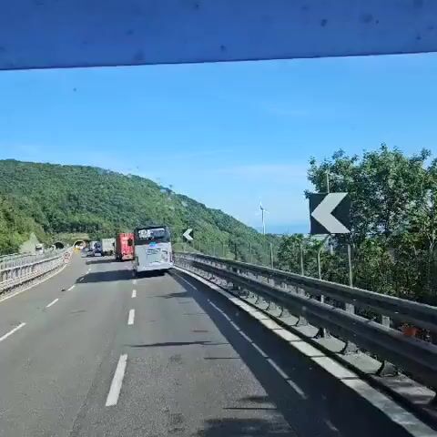 The video shows a vehicle traveling on a highway alongside a mountain range with green trees. The sky is blue and clear. There is a tall bridge in the distance crossing a valley and a wind turbine on a hill in the background. A bus is also driving along the highway.