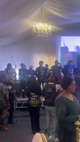 A band is performing on a small stage in front of an audience. The band consists of a saxophone player in the center, a singer on the right, a keyboard player behind the saxophone player, and female singers on the left. There is a large chandelier hanging overhead and white curtains behind the band. A TVC camera crew is filming the performance from the front. The audience includes people wearing shirts that say "XRO SOUL" and "I think Growth Think DM360 Degree". A large screen showing a woman is visible in the background. The room is brightly lit with a blue hue.
