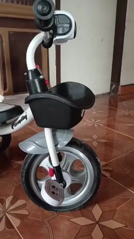 A white and black child's tricycle is shown in a room with tiled flooring. The tricycle has a black basket at the front, a grey fender over the front wheel, and a grey seat with a black backrest. The word "Family" is visible on the side of the tricycle. The camera pans across the tricycle, showing it from different angles.