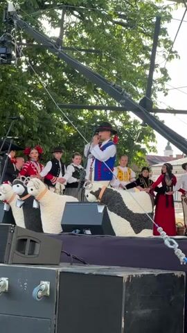A group of children and adults in traditional folk costumes are performing on a stage outdoors. The performers are standing behind two large, white, fluffy objects that resemble sheep or lambs, decorated with black and white floral patterns. A young boy in the center is singing into a microphone, wearing a blue vest over a white shirt and a black hat. Other children are dressed in red and white outfits with vests and skirts, or vests and pants, some are holding instruments like violins. A woman in a red dress stands to the right of the stage. The stage appears to be set up outdoors with trees in the background and a large camera crane overhead. The scene is brightly lit, suggesting it is daytime.