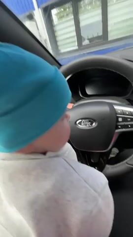 A baby with light skin and wearing a light blue beanie and gray hooded sweatshirt is sitting in the driver's seat of a car. The baby is facing forward, looking at the steering wheel and the car dashboard. The baby reaches out with one hand and touches the center of the steering wheel. The car's interior is dark with a black leather steering wheel and a digital dashboard. The car appears to be at a car wash.
