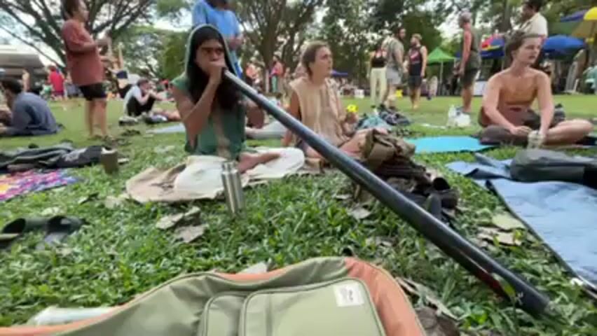 A wide shot shows a group of people sitting on the grass at a park or outdoor gathering. A person wearing a headwrap and a green top sits cross-legged and plays a long, black tube instrument. Two women sit near the musician, seemingly listening. Others are visible in the background, some standing and walking around. Several bags, blankets, and personal items are scattered on the grass around the seated individuals. A guitar case sits in the foreground. The scene appears to be relaxed and informal.