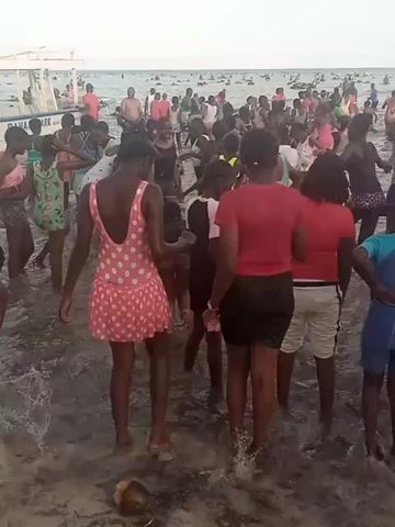 A crowded beach scene features a large group of people wading in shallow water. Most appear to be children and teenagers. They are dressed in various swimsuits and casual clothing. Some are splashing and playing. A white motorboat is partially visible in the background, close to the shoreline. In the distance, the ocean is filled with more people, suggesting a popular beach location. The sky is overcast, and the overall lighting is somewhat muted. The sand is dark.