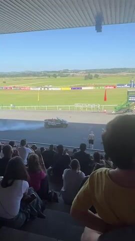 The video captures a crowd of spectators seated on bleachers, looking out at a race track. In the center of the track, a car is performing a drift, emitting a large plume of smoke. Two individuals are standing near the car. Beyond the track, a large, green field stretches out under a bright blue sky, with rolling hills in the distance. The scene appears to be a daytime event at a racing venue.