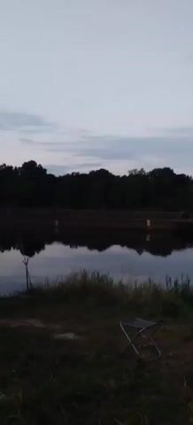 A barge is moving on a river. The sky is overcast. There are trees on the opposite bank. There is grass and a folding chair on the near bank.